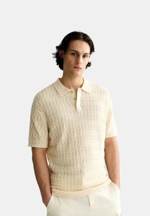 Junger Mann mit dunklen Haaren, der ein cremefarbenes kurzärmeliges, strukturiertes Poloshirt und helle Hose trägt, steht mit den Händen in den Taschen.