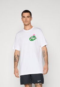 Camiseta blanca de algodón con un gráfico floral colorido en el pecho; presenta un corte relajado y mangas cortas. El modelo lleva pantalones cortos negros.