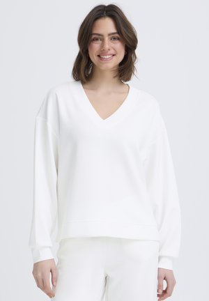 Giovane donna con capelli castani a lunghezza spalla, sorridente, indossa una maglia bianca a maniche lunghe con scollo a V ampia e pantaloni bianchi abbinati.