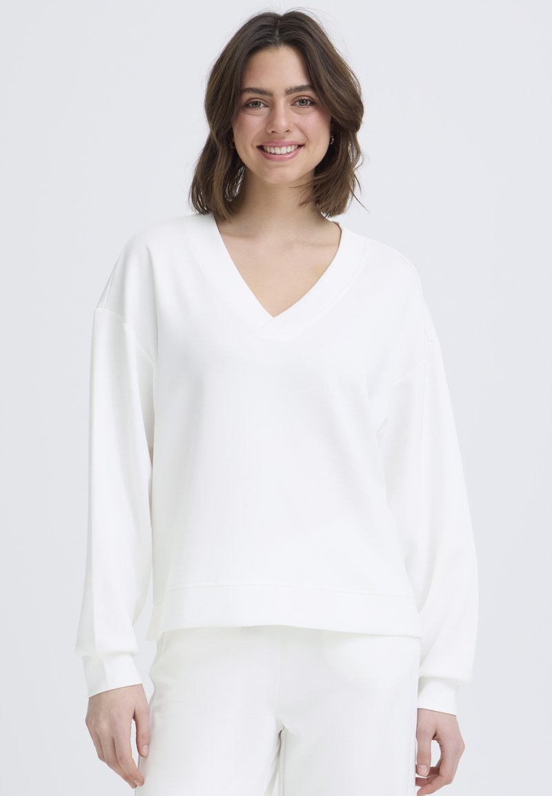 Mujer joven con cabello castaño hasta los hombros, sonriendo, vistiendo una blusa blanca suelta de manga larga con cuello en V y pantalones blancos a juego.