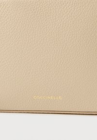 Smėlio spalvos odinė rankinė su tekstūruota paviršiaus danga ir subtiliu blizgesiu. Auksinė įspausta „Coccinelle“ logotipo žyma apatiniame dešiniajame kampe. Elegantiškas ir minimalus dizainas.