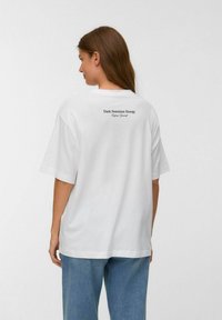 Witte katoenen T-shirt met een ruime pasvorm, korte mouwen en een grafische print op de rug met de tekst "Donkere Vrouwelijke Energie – Laat Jezelf Zien."