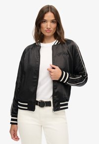 Vrouw in een zwarte satijnen bomberjack met witte strepen, witte shirt, witte broek en zwarte riem, staand tegen een effen achtergrond.