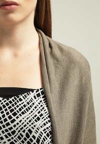 Brązowy dziergany szal opada na ramiona, z teksturowanym wzorem. Pod spodem widoczna jest czarna bluzka z białym, abstrakcyjnym wzorem kratki.