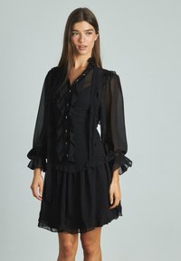 Abito nero con accenti arricciati, apertura frontale con bottoni e maniche lunghe trasparenti. Tessuto fluido con dettagli a strati e orlo frastagliato.