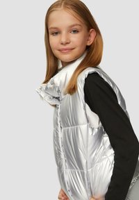 Zilveren geïsoleerde vest met drukknoopsluiting, hoge kraag en opgeblazen secties. Draag het over een zwart langemouwen shirt. Glad, glanzende stofafwerking.