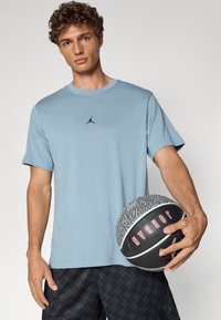 Lys blå bomulls t-skjorte med rund hals og svart logo, iført sorte mønstrede shorts, holder en basketball med teksturert overflate.