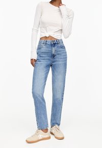 Lehký bílý top s dlouhým rukávem a uzlem na spodním lemu, kombinovaný s vysokým pasem mírně vyšisovaných modrých džínů a b�éžových tenisek s bílými detaily.