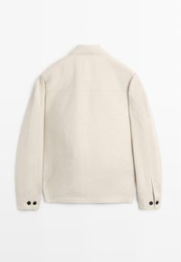 Beige langärmelige Jacke mit geradem Kragen und schwarzen, mit Knöpfen versehenen Manschetten, von hinten auf weißem Hintergrund gezeigt.