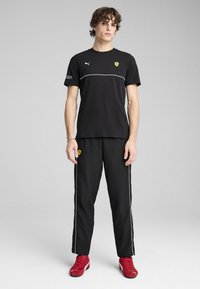 Sort atletisk t-shirt med hvid accentlinje, Puma-logo og gul Ferrari-emblem; parret med sorte bukser med sidelommer. Røde sko.
