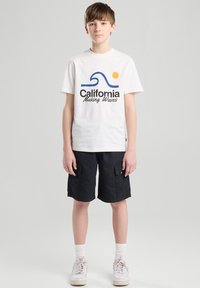 Valkoinen puuvillapaita, jossa on sininen aalto-design ja "California Making Waves" -teksti. Mustat cargo-shortsit. Mallilla on päällään valkoiset lenkkarit ja nilkkasukat.