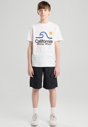 Valkoinen puuvillapaita, jossa on sininen aalto-design ja "California Making Waves" -teksti. Mustat cargo-shortsit. Mallilla on päällään valkoiset lenkkarit ja nilkkasukat.