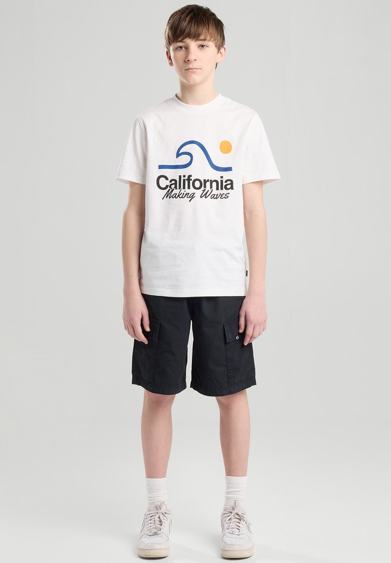 Valkoinen puuvillapaita, jossa on sininen aalto-design ja "California Making Waves" -teksti. Mustat cargo-shortsit. Mallilla on päällään valkoiset lenkkarit ja nilkkasukat.