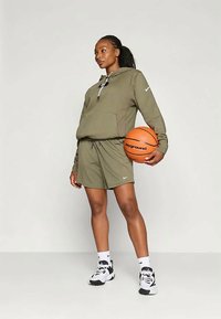 Hanorac Nike de culoare verde măslinie cu sfoară, pantaloni scurți asortați și adidași Nike negri. Ținând o minge de baschet, stând pe un fundal simplu.