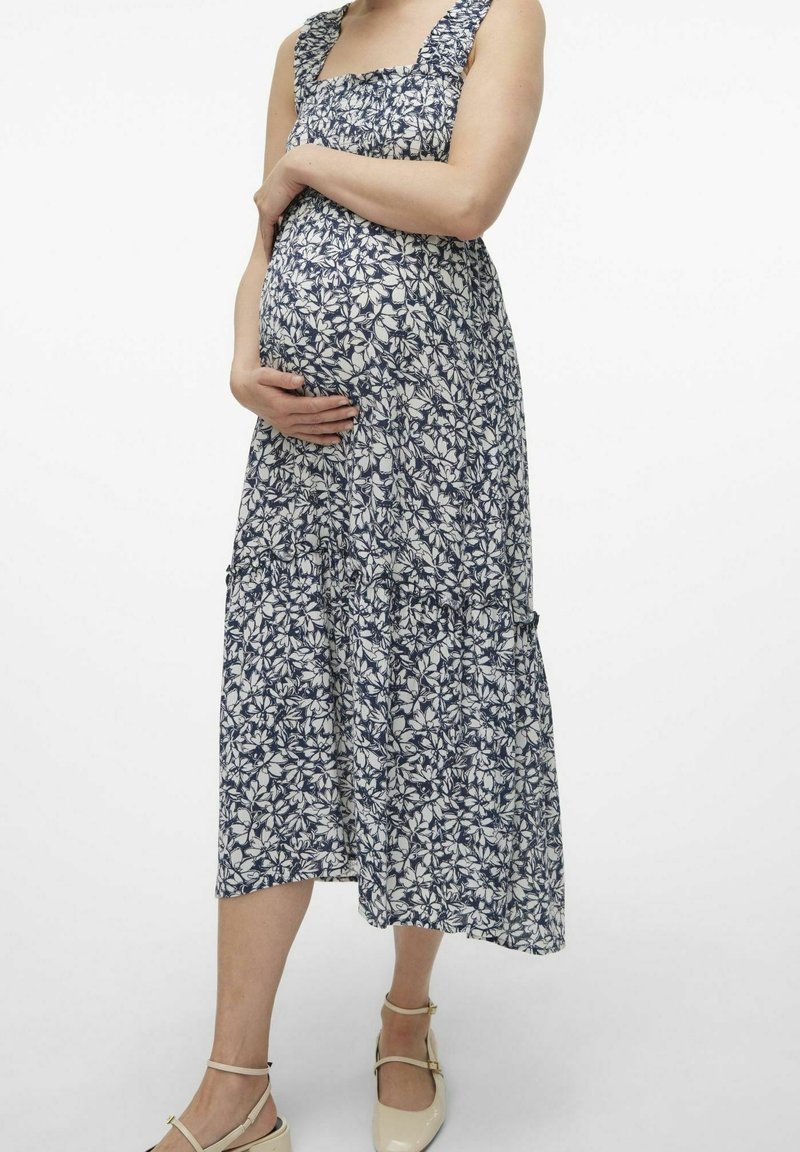 Femme enceinte en robe fleurie sans manches, tenant son ventre, portant des ballerines beiges à bride cheville, debout devant un fond uni.