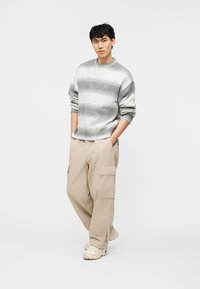 Νεαρός άνδρας που στέκεται με τα χέρια στις τσέπες, φορώντας γκρι ριγέ πουλόβερ, μπεζ παντελόνι καρό και εκρού αθλητικά σε λευκό φόντο.