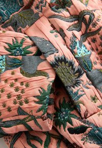 Gewatteerde textiel met een koraalrode achtergrond en groene bladeren en bloemenborduurwerk. Gestructureerde stof met gevarieerde stikpatronen.