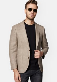 Beżowy żakiet w jodełkę z dwoma br�ązowymi guzikami, noszony na czarnej koszulce i ciemnych dżinsach. Modelka pozuje w okularach przeciwsłonecznych, z rękami w kieszeniach.