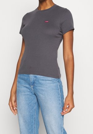 Femme portant un t-shirt Levi's gris foncé ajusté et un jean en denim bleu clair, debout devant un fond clair uni.
