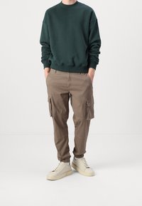 Persoon in een donkergroene sweatshirt, bruine cargobroek met zakken en off-white sneakers, staande met handen in de zakken.