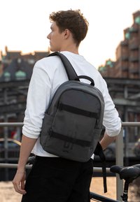 Šedý batoh s nastaviteľnými popruhmi, plochým predným dielom a dvomi prackami. Hladký povrch a minimalistický dizajn, ktorý nosí osoba v bielom tričku.