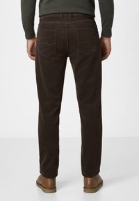 Mann in dunkelbraunen Cordhosen, braunem Gürtel, dunkelgrünem Langarmhemd und braunen Leder-Schuhen, steht vor einem schlichten Hintergrund.