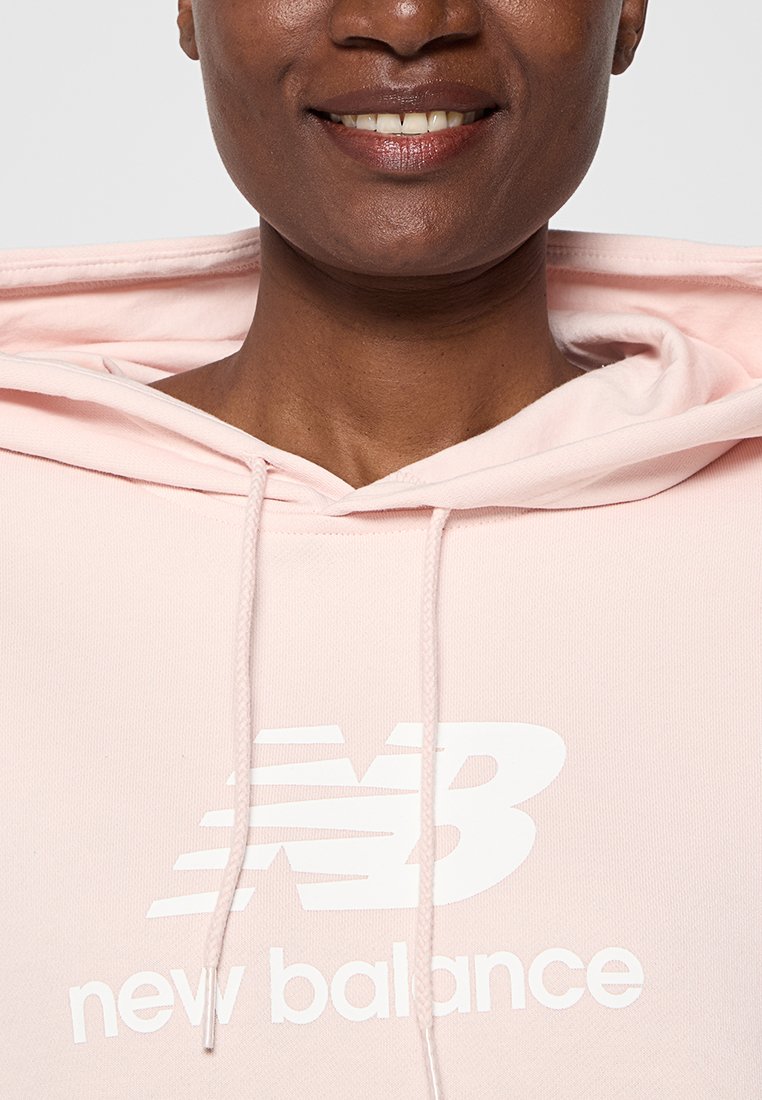 Sudadera rosa claro hecha de una tela suave, con un logotipo blanco de "New Balance" en el frente y cordones en la capucha.