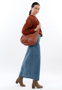 Bruine leren schoudertas met een gebogen vorm, voorzien van een rits en een reliëflogo. Model draagt een roestkleurige gebreide trui en een denim rok.