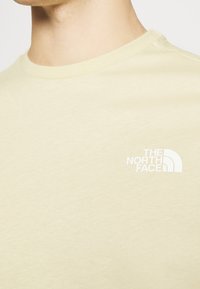 Primer plano de una persona que lleva una camiseta beige con un logo blanco de "The North Face" en el pecho.