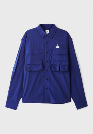 Blaues langärmeliges Hemd mit zwei Brusttaschen, Knopfleiste und einem dreieckigen Logo auf der linken Seite. Aus leichtem Stoff gefertigt.