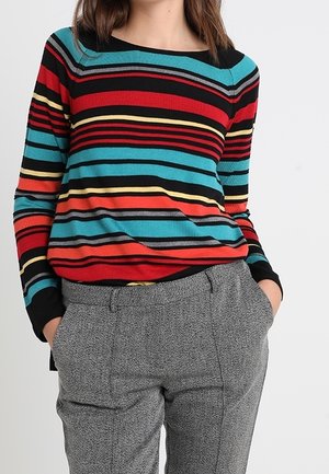 Femme portant un t-shirt à manches longues à rayures horizontales colorées et un pantalon gris texturé, les mains dans les poches, sur un fond uni.