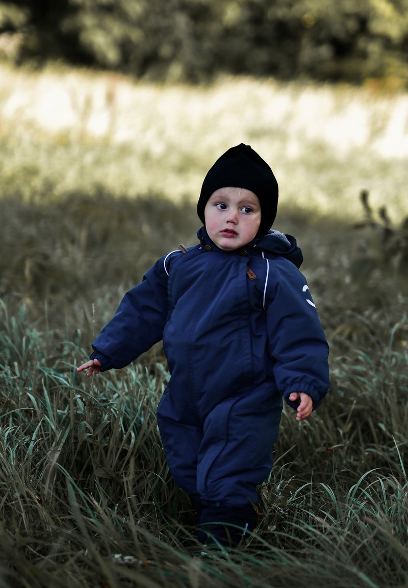 Niño con un mono de nieve azul oscuro con aislamiento y detalles en blanco, llevando un gorro de punto negro, caminando entre hierba alta.