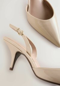 Smėlio spalvos lakuotos odos aukštakulniai bateliai su slingback dirželiais, smailiomis nosytėmis ir reguliuojamomis sagtelėmis, padėti ant baltos paviršiaus.