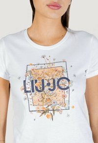 Camiseta blanca de algodón con estampado floral y el texto "LIU•JO" en azul oscuro. Cuello redondo, mangas cortas y detalles cuidadosamente elaborados.
