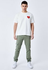 Hvid kortærmet t-shirt med et rødt brudt hjerte grafisk design, parret med grønne cargo bukser med sidelommer og hvide sneakers.