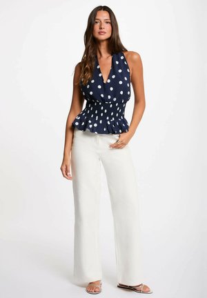 Donna che indossa un top smanicato blu scuro a pois con vita arricciata e pantaloni bianchi a gamba larga, in piedi con una mano sul fianco.