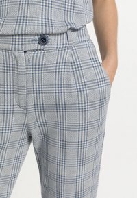 Person wearing blue and white plaid trousers with a buttoned waistband, one hand casually in pocket against a white background.