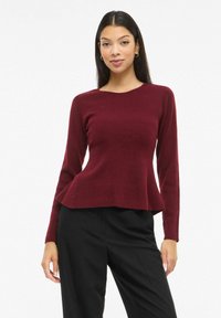 Tumepunane soonikkoes pikkade varrukatega kampsun, millel on lõhikuga hem, valmistatud pehmest materjalist. Sobitatud mustade laiade pükstega.