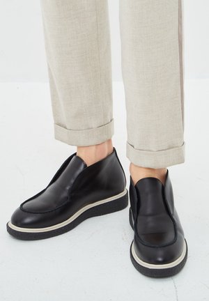Schwarze Leder-Slipper mit dicken weißen Sohlen, getragen zu beigen, aufgekrempelten Hosen, auf weißem Boden.