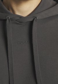 Gros plan sur un sweat à capuche gris foncé avec des cordons épais et le logo "HUGO" en relief centré sous l'encolure.