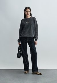 Jeune femme debout tenant un petit sac noir, portant un sweat-shirt Off-White gris foncé, un pantalon noir orné de petits clous, et des chaussures à imprimé léopard.