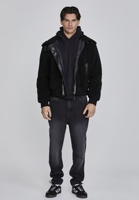 Chaqueta negra de piel sintética de borreguillo con detalles de cuero, combinada con una sudadera negra con capucha, jeans oscuros y zapatillas negras; diseño sencillo y minimalista.