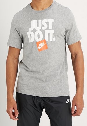 Mies, jolla on harmaa Nike "JUST DO IT." T-paita ja mustat Nike shortsit, seisoo käsivarret rentoina sivuilla.