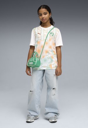 Giovane donna che indossa una T-shirt bianca con motivo, jeans azzurri strappati, sneaker argento e una piccola borsa a tracolla verde Puma.