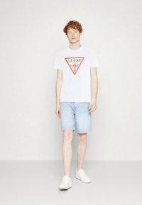 Camiseta blanca con logo triangular rojo, combinada con pantalones cortos de mezclilla azul claro y zapatillas blancas. El modelo está de pie frente a un fondo liso.