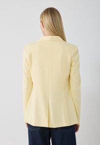Blaser jaune ajusté avec une texture lisse, épaules structurées et une fente simple à l'arrière, associé à un pantalon en denim foncé ample.