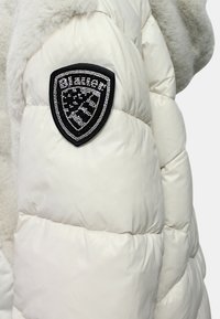 Chaqueta blanca acolchada con una textura suave y un collar parecido al de piel, que presenta un parche negro en forma de escudo con piedras brillantes en la manga.