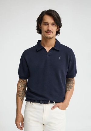 Man met halflang donker haar en tatoeages op beide armen, draagt een marineblauw poloshirt en witte broek, staand tegen een effen achtergrond.