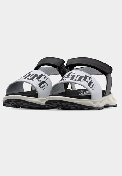 Ανοιχτά σανδάλια ασπρόμαυρα με χοντρές σόλες, ρυθμιζόμενους μαύρους ιμάντες και έντονους λευκούς ιμάντες με μαύρα τυπωμένα γράμματα.