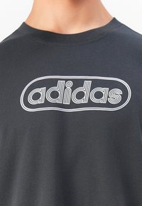 Κοντινός πλάνο ενός σκούρου γκρι μπλουζάκι με το λογότυπο "adidas" σε λευκή περιγράμματος στο στήθος.